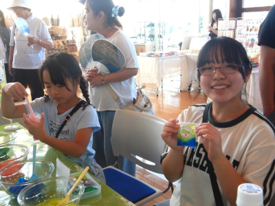 作品を手に持ち笑顔でカメラを見つめる女の子と、隣で保冷剤作りに取り組んでいる女の子の写真。