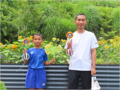 ひまわり畑の前で、手作りのフォトプロップスを手に持って並ぶ男の子と男性の写真