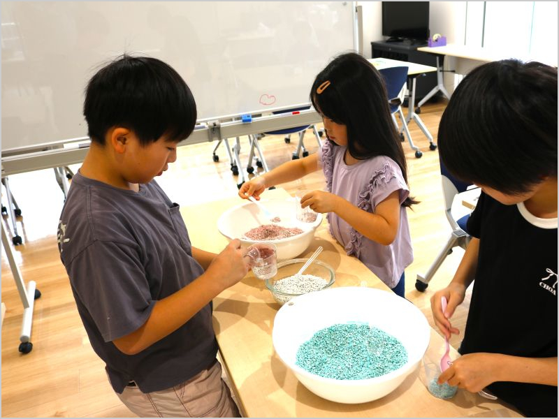 スプーンで透明なカップにすくい入れている3人の子どもたちの様子